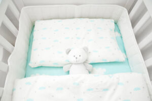 Small, white teddy bear in baby bed. Linen with clouds. Top view.