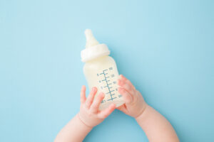Infant hands holding bottle of milk on light blue floor background. Feeding time. Pastel color. Closeup. Point of view shot. Top down view.