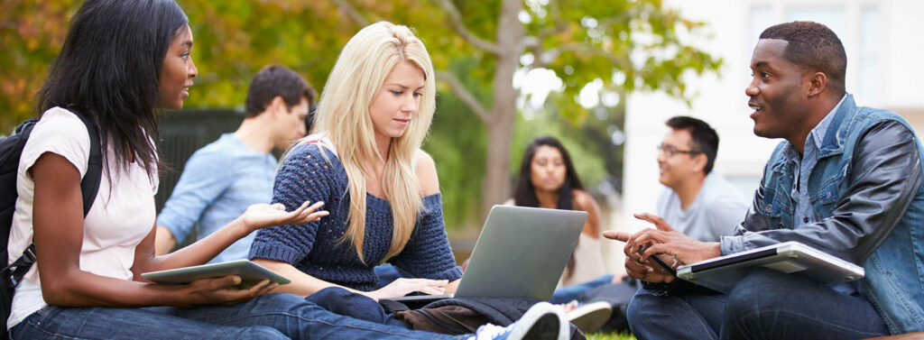 Three adults with their laptops having a conversation outside. Three adults in the background conversing.