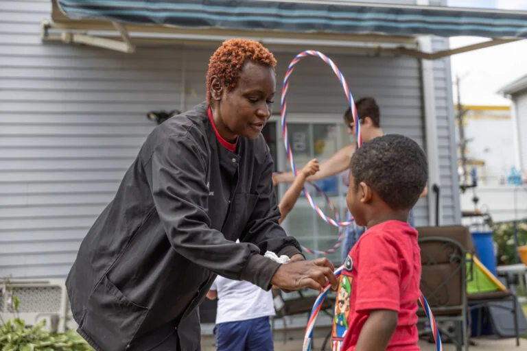 A women and a child at a house playing hula hoop at a house. A teenager and child are playing hula hoop in the background.