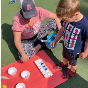 Child and educator outside on turf doing arts and crafts.